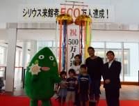 「シリウス来館者500万人達成」と書かれた幕が掲げられ、割られたくす玉の下で記念撮影をしているヤマトン、河原さんご一家と市長の写真