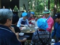 大きな炊き出し用の鍋をお玉でかき混ぜている女性と配膳の順番を待って並んでいる人の写真