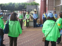 黄緑のジャンパーを着た方々が集まり、反射ベストを着た男性が話をする写真