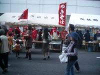 焼きそばと書かれた旗が立つテント内で、赤い服を着た人々が焼きそばを焼き、その前にいる通行人の写真