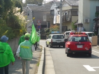 赤いパトロールカーの横を、緑のベストと帽子を被り、旗をもって歩いている人々の写真