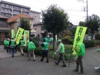 防犯パトロールと書かれたのぼり旗を持ち、緑の上着を着た人たちが縦に並んで歩いている写真