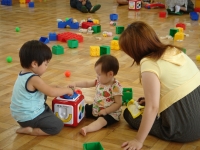 女性が見守るなか、2名の幼児がおもちゃで遊んでいる子育てサロンの様子の写真