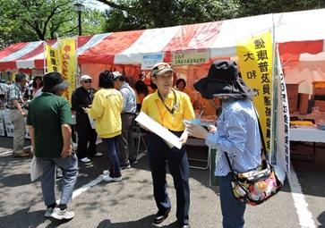 イベント会場のテント前で黄色いポロシャツ着た健康普及員が、来場者に資料を配布している写真