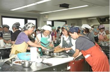 調理室で三角巾とエプロン姿の人々がグループごとに分かれて料理をしている写真