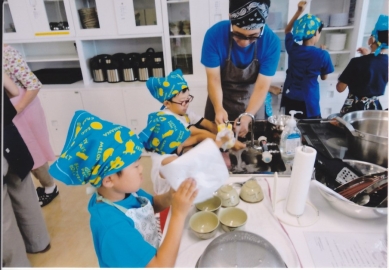 お揃いの水色に黄色の模様が入った三角巾をした子供達と一緒に料理をしているお父さんの写真