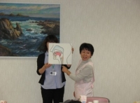 左の女性が人の口から喉にかけて描いているイラストを持ち、右側のピンク色のエプロンを着用した女性が説明している写真