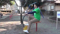 男性が公園に設置された健康遊具クルクルサイクルの椅子に腰かけ、バーにつかまってペダルをこいでいる写真