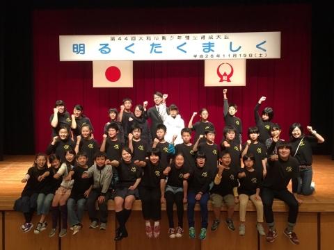黒いTシャツを着た児童たちが舞台に腰を掛け、拳を上に挙げて記念撮影している写真