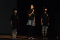 黒いTシャツを着た児童3人が舞台に並んでおり、真ん中の女子児童がマイクを持って発表している写真