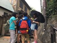 建物の脇の階段下で黒色の傘をさしている女の子が話しをしているのを他のメンバーが聞いている写真