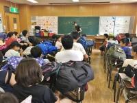 夏の宿泊研修参加の子供達が各机に座り黒板前の担当者の人の話を聞いている写真