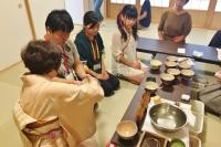 3名の女の子が長机の前に座り、手前の着物を着た女性から茶道の作法を教わっている写真