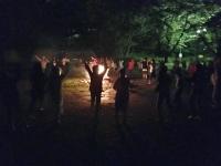 参加者の人達の円の中心にキャンプファイヤーの火が灯されている夜の暗闇の中の写真