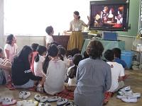 大きなテレビの左側に立つ女性が映し出された映像の説明をしているのを、前に座っている子供たちが聞いている写真