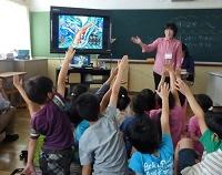 教室の黒板の前に置かれた大きなテレビの右側に立つ女性と、床に座って手を挙げている子供たちの写真