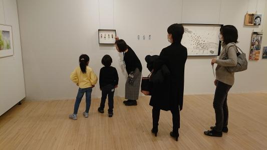 額に入った作品に興味津々の2名の子どもが、作品の説明をしている女性の話しを聞いている写真