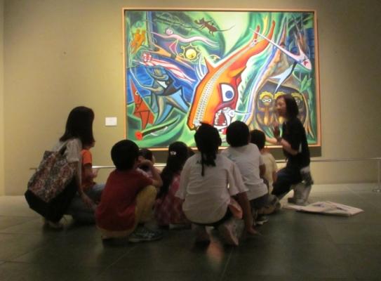 美術館内の大きな絵画の前に子ども達が座って右側の女性の説明を聞いている写真