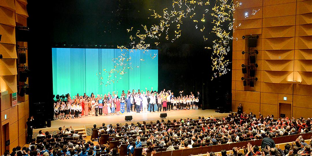 舞台上の多くの方達に、ホールに集まった多くの方達が拍手を送っていいる画像