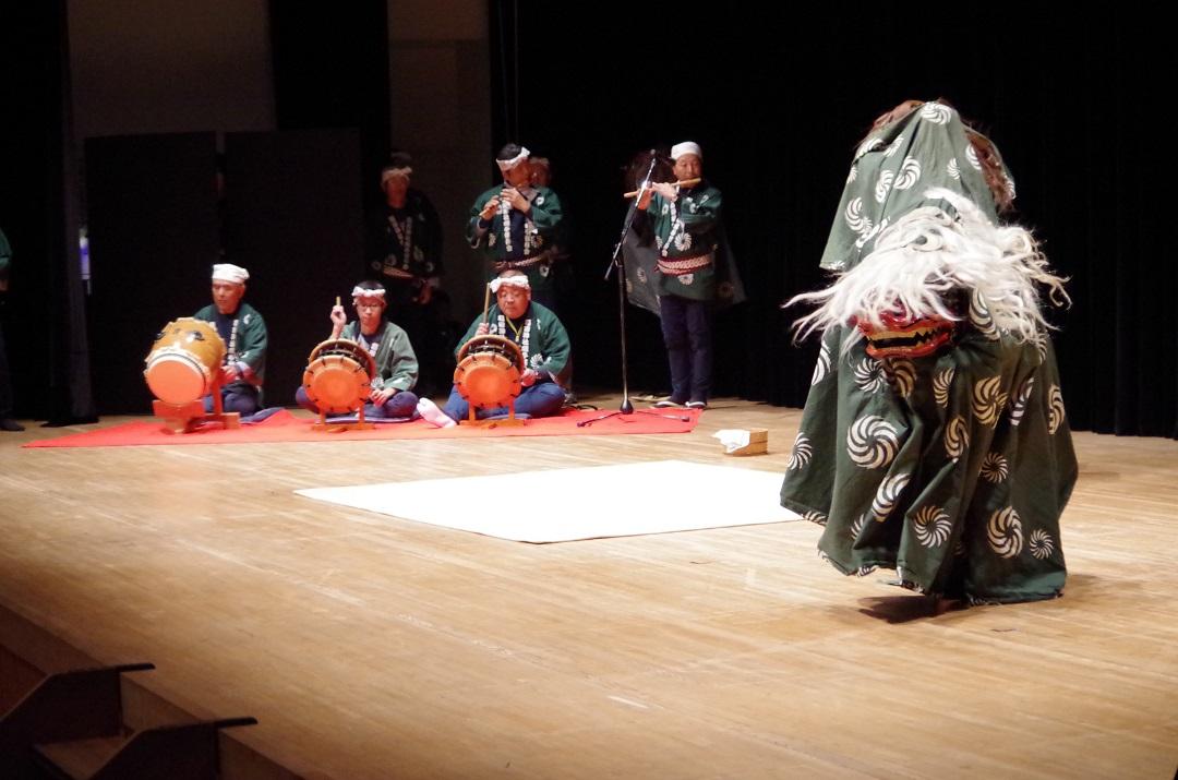 舞台の上で太鼓や笛を演奏している男性と囃子に合わせて踊る獅子舞いの写真