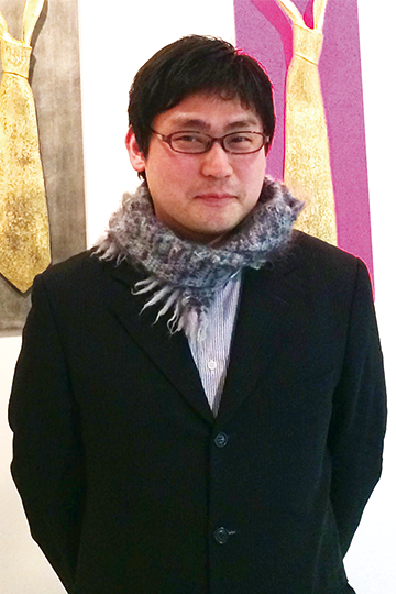 紺色のジャケットを着用し首にマフラーを巻いた大塚 亨さんが作品の前に立っている写真