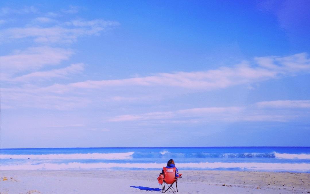 白い雲が浮かんでいる青い空と青い海の見える砂浜に,赤い椅子に座った女性が海を眺めている後ろ姿の写真
