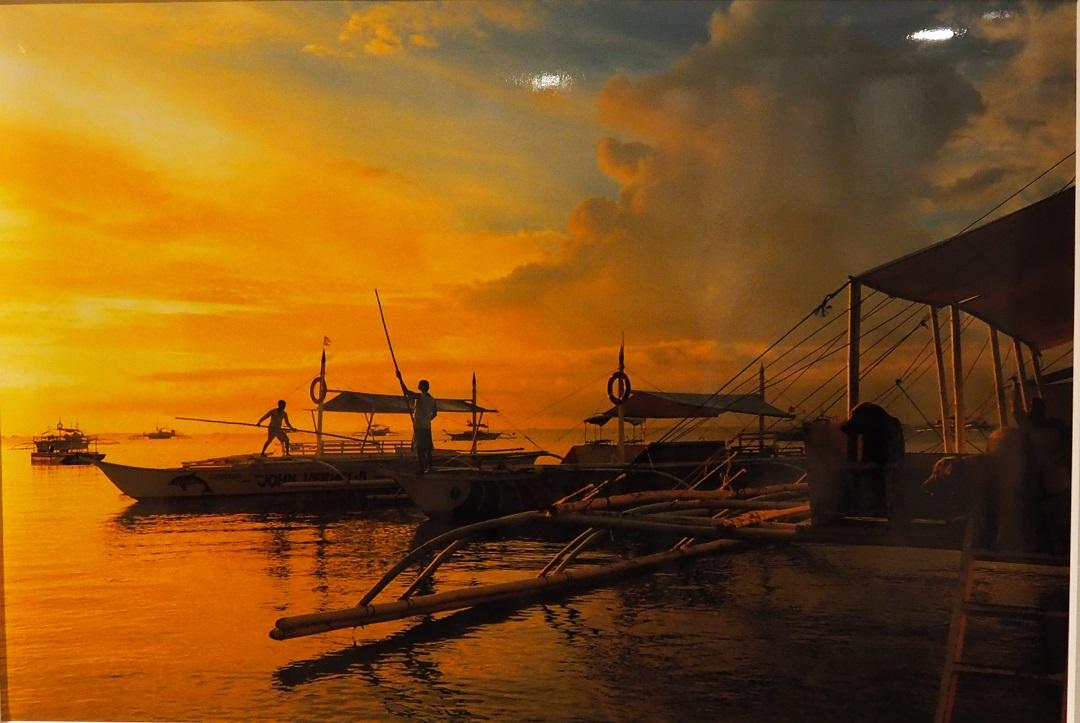 オレンジ色に染まった夕暮れ時の海で、2人の男性が長い棒をもち各自の船に乗っている様子を港で2匹の犬が待っている写真