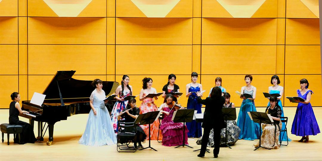 グランドピアノの横で、鮮やかな色や柄のドレスを着た女性が楽器を演奏したり、合唱をしている写真