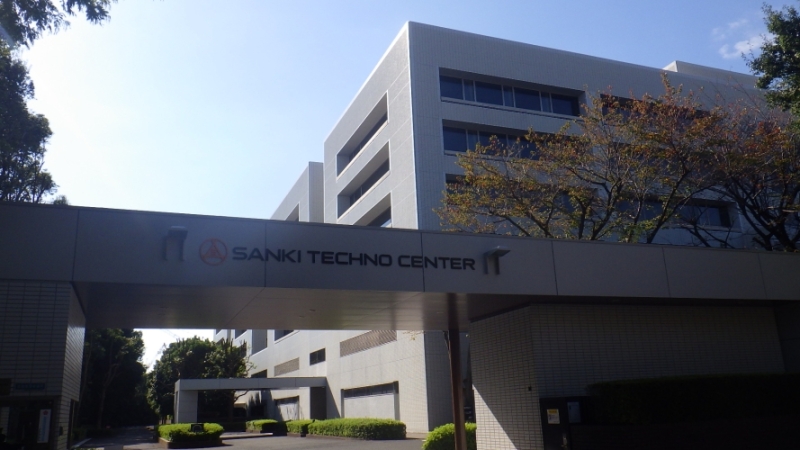 手前に「SANKI TECHNO CENTER」と書かれた門のような入口があり、奥に四角い大きな建物が見える三機テクノセンターの外観写真