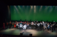舞台上の奥に赤い丸のようなものを持った人たちが並んでいて、中央で一人の白いジャケット姿の男性が立っている写真