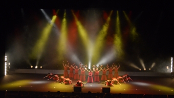 舞台上で赤い衣装を着た女性たちが両手を上にあげたり、しゃがんだりしながら踊っている写真