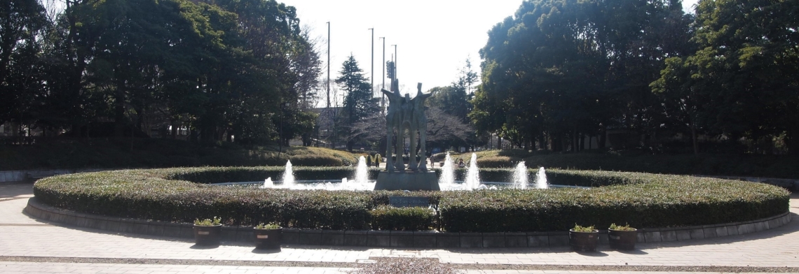 丸く形作られた噴水、中央には人間のような形をした三体の銅像が立っている公園の写真