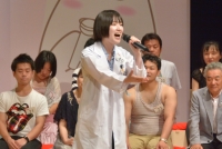 奥には出場者たちが座っていて、手前には白衣を着た女性が手を前に差し出しながら歌っている写真