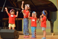 四人おそろいのオレンジのTシャツを着た子供たちが舞台上で手を上に挙げながら歌っている写真