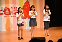虹の絵が描かれたおそろいのTシャツを着た3名の女の子がマイクを握りしめ歌っている写真