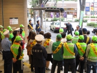 交通安全と書かれたタスキをかけ、緑の帽子とベストを着た人たちが集まっている様子の写真