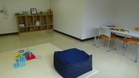 奥に棚、右側に椅子とテーブル、中央に紺色のクッションが置かれている教育支援教室「ひだまりの教室」の室内写真